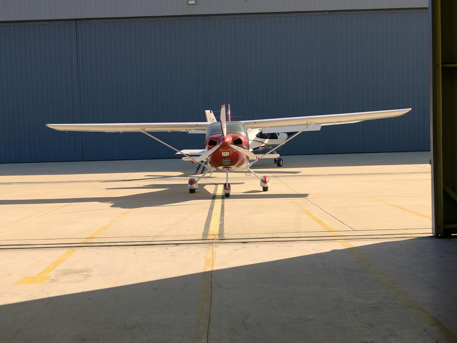 A white high-wing training airplane parked in the open doorway of a hangar on a sunny day